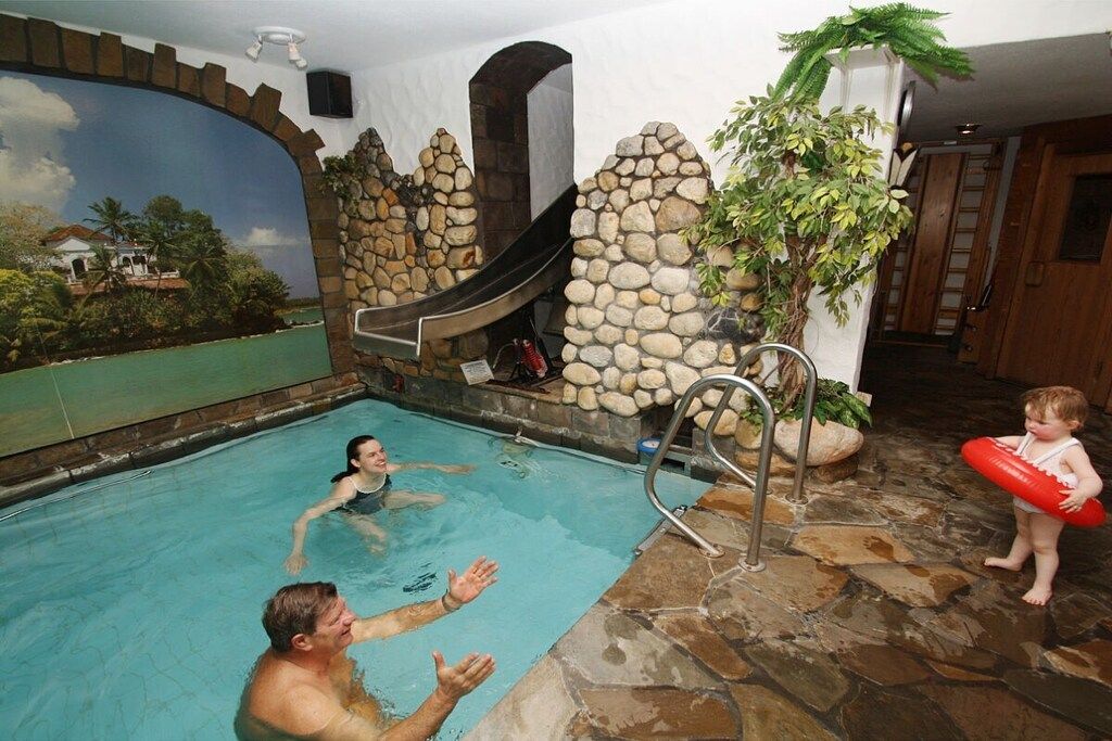 An indoor swimming pool with a stone wall and a slide, a child with a floatation device, and a person in the water.