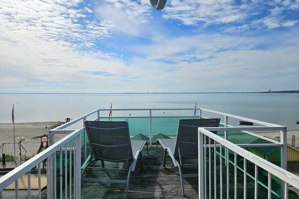 Eine Terrasse mit Stühlen, die auf ruhiges Gewässer blickt, mit Geländer und blauem Himmel.