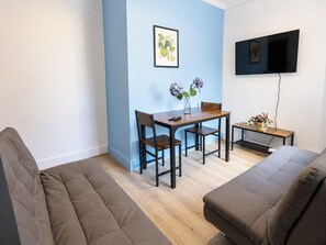 A modern living room with a sofa, a dining table with chairs, a small side table with a plant, and a flat-screen TV mounted on the wall. - Central 4-BR Middlesbrough | Walk to Station & Uni (Middlesbrough)