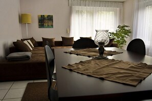 Een woonkamer met een bank, een eettafel met placematen, een lamp en een schilderij aan de muur.