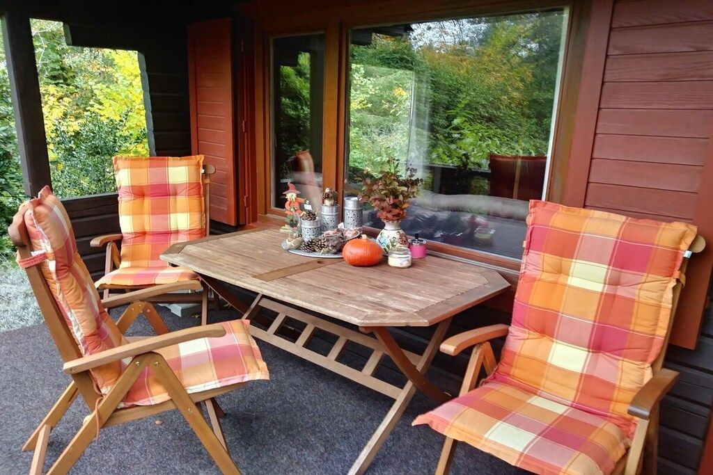 A wooden table with chairs, a pumpkin, and decorative items on a porch with a view of trees.