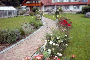Un chemin menant à une maison avec un toit rouge, entourée d’un jardin où poussent différentes plantes et fleurs.