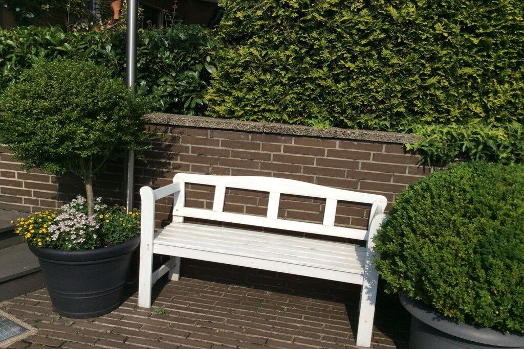 A white bench in front of a brick wall with a hedge and potted plants.
