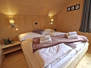 A wooden cabin bedroom with a double bed, bedside lamps, a shelf with a book and a clock, and a curtain on the window. - Cabin in Hohentauern With Sauna (Hohentauern)