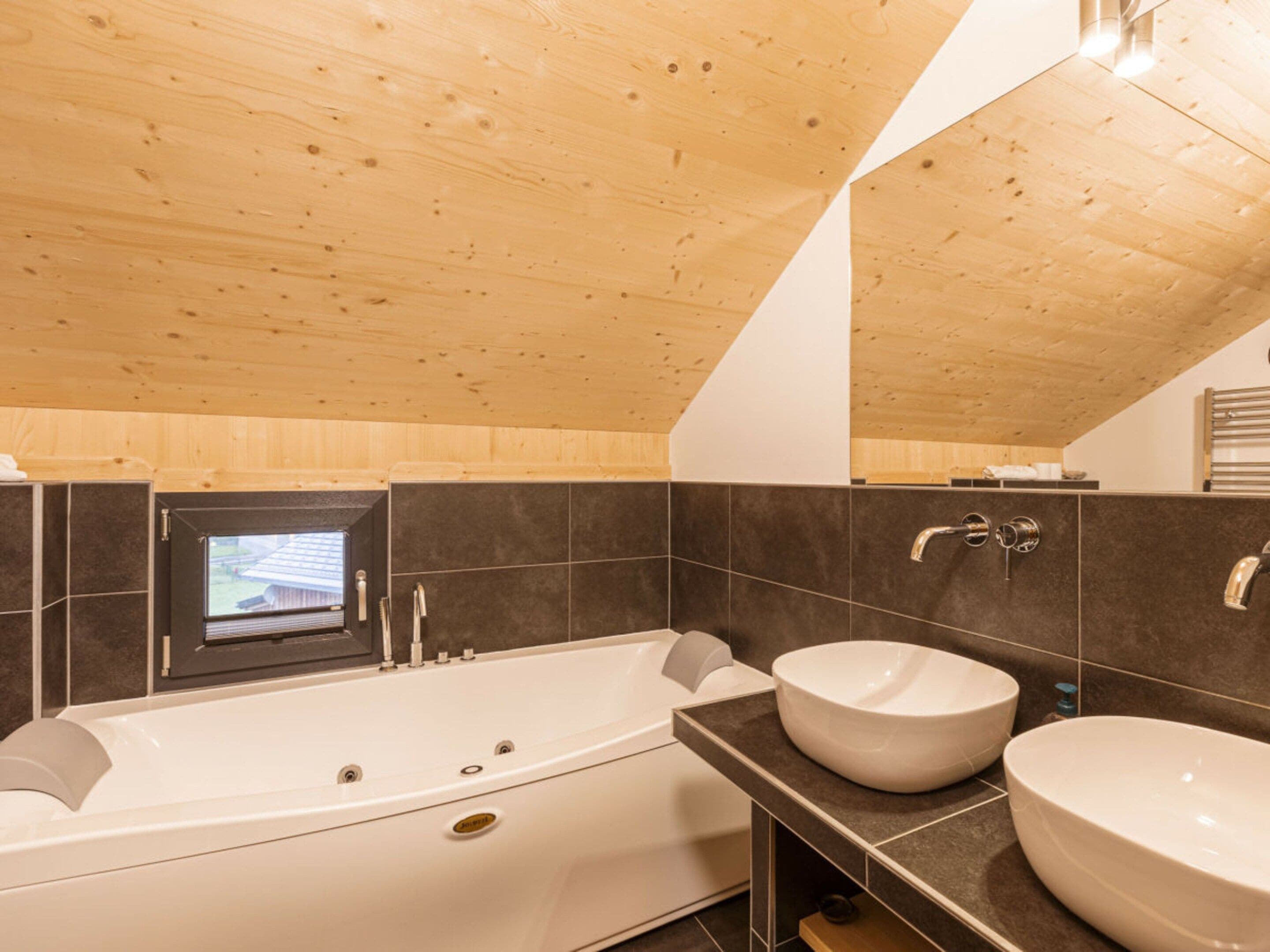 Ein modernes Badezimmer mit Holzboden, einer großen Badewanne, zwei Waschbecken und einem kleinen Fenster.
