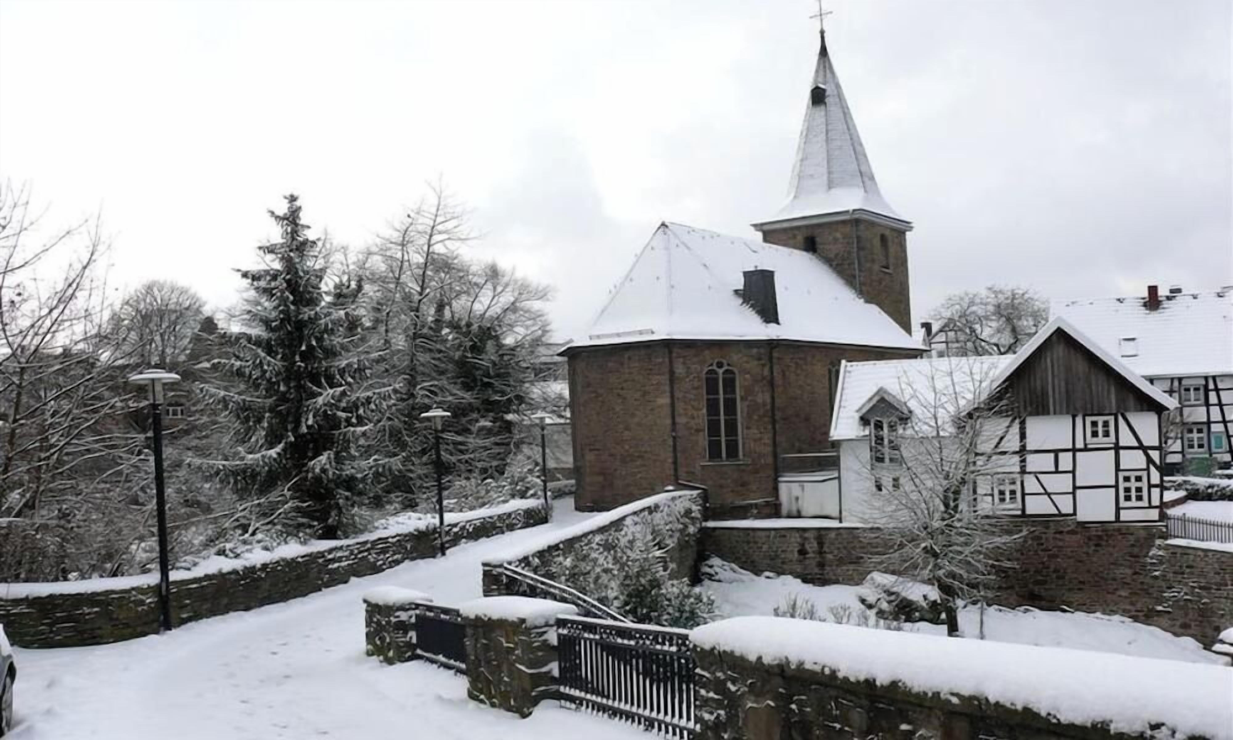 A church with a steeple, a stone wall, and snow-covered trees.
