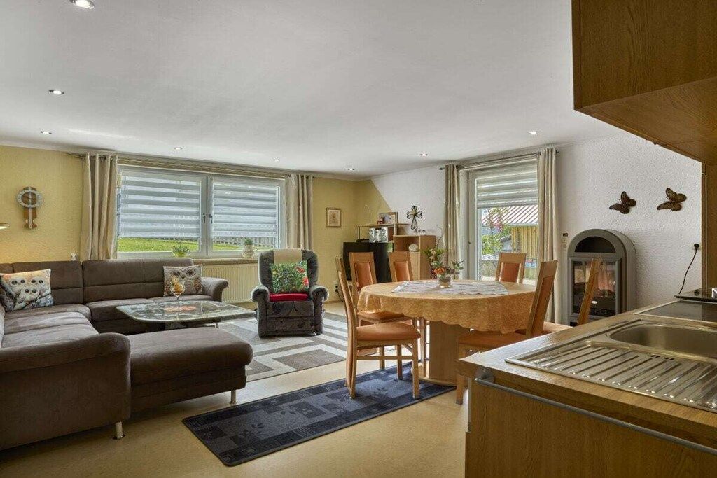 A living room with a sofa, armchair, and dining table. There is a fireplace and a window with blinds.