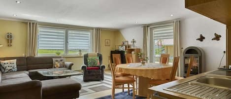 Una sala de estar con sofá, sillón y mesa de comedor. Hay una chimenea y una ventana con persianas.