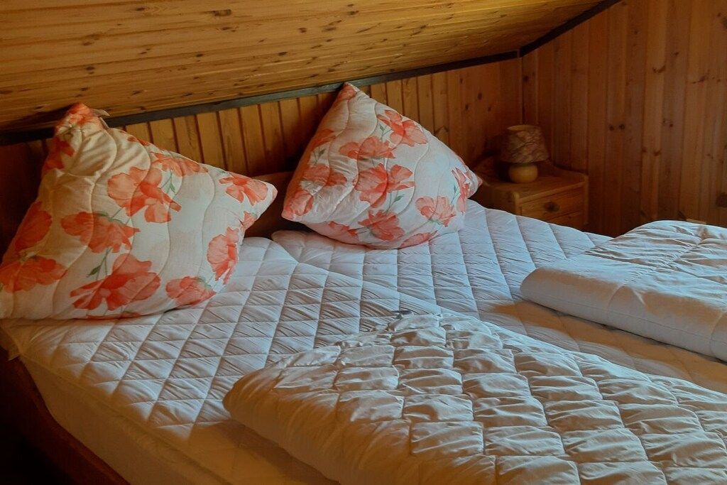 Una cama con edredón blanco acolchado y dos almohadas florales, frente a una pared de madera en una habitación con techo inclinado.