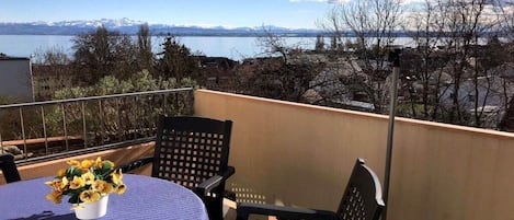 A balcony with a table and chairs, a vase of flowers, and a view of a lake and mountains.