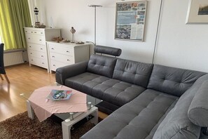 A modern living room with a grey sofa, a small table with a pink tablecloth, and a white dresser. - Döse beach high-rise (Cuxhaven)