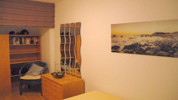 A bedroom with a bed, a wooden dresser, a chair, a bookshelf, and a wall-mounted mirror. - Döse beach high-rise (Cuxhaven)