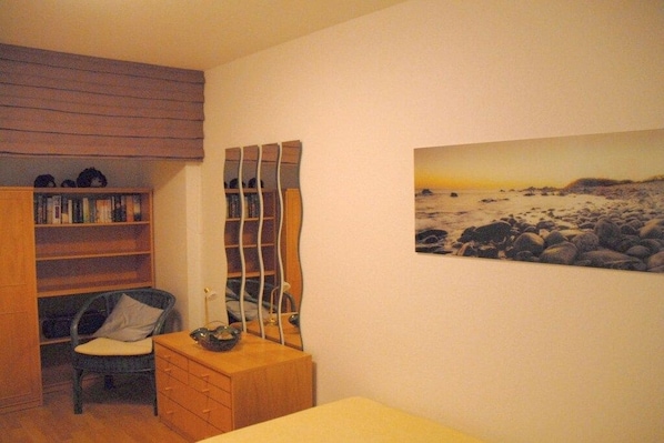 Quarto com cama, cômoda de madeira, cadeira, estante e espelho fixado na parede.
