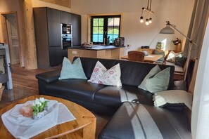 A modern living room with a black leather sofa, a wooden coffee table, and a kitchen area with a built-in oven and sink. - Raccoon Hut Steirer-Hof (Vöhl)