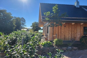A wooden house with a sloped roof, surrounded by a garden with various plants and a stone wall. - Raccoon Hut Steirer-Hof (Vöhl)