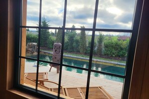 Blick durch ein Fenster auf eine Holzterrasse, einen Pool und einen Stuhl.