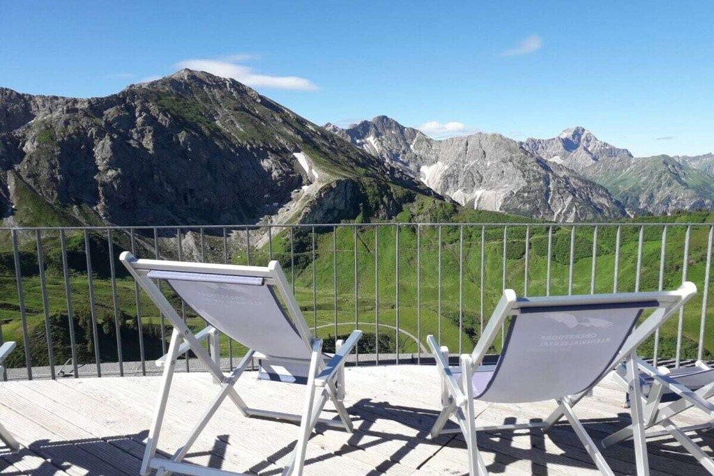 Ein Balkon mit zwei weißen Klappstühlen, der auf eine gebirgige Landschaft blickt.