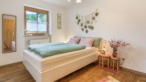 A bedroom with a bed, a window, a wall-mounted plant, a bedside table with a lamp, and a mirror.