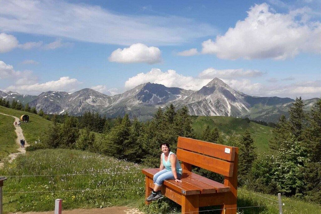 Eine Person sitzt auf einer Bank, im Hintergrund eine gebirgige Landschaft.
