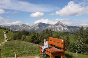 Eine Person sitzt auf einer Bank, im Hintergrund eine gebirgige Landschaft.