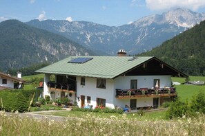 Eine zweistöckige Chalet mit grünem Dach, Balkonen mit Blumenkästen und Bergblick.