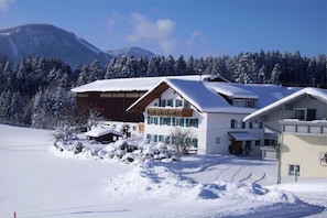 Un bâtiment à deux étages, dont le toit est recouvert de neige, entouré d’un paysage enneigé et d’une forêt.