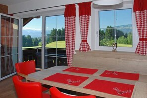Ein Zimmer mit einem Esstisch, roten Stühlen und Blick auf eine grüne Landschaft durch große Fenster.