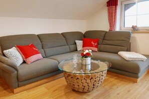 Un salon avec un canapé gris, des coussins à carreaux rouges et blancs, une table basse en verre avec un pied en bois, et une fenêtre ornée de rideaux à carreaux rouges et blancs.