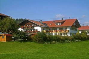 Ein zweistöckiges Haus mit rotem Dach, einem Balkon mit roten Blumen und einer kleinen Holzstruktur zur Linke.
