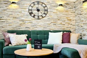 A green sofa with cushions, a round wooden table with a vase and a sign, and a wall clock. - Holiday apartment with 1 bedroom (Garmisch-Partenkirchen)