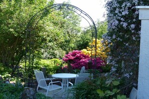 Ein Garten mit einem weißen Metalltisch und Stühlen, umgeben von üppigem Grün und blühenden Pflanzen.