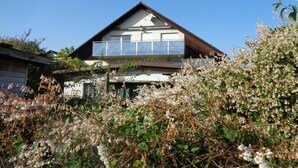 Una casa a due piani con un balcone, circondata da vegetazione rigogliosa.