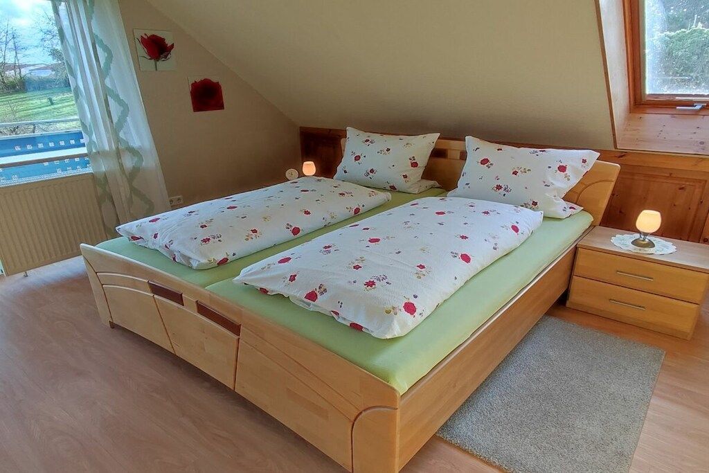 A double bed with floral bedding, a bedside table with a lamp, and a view of the outdoors through a window.
