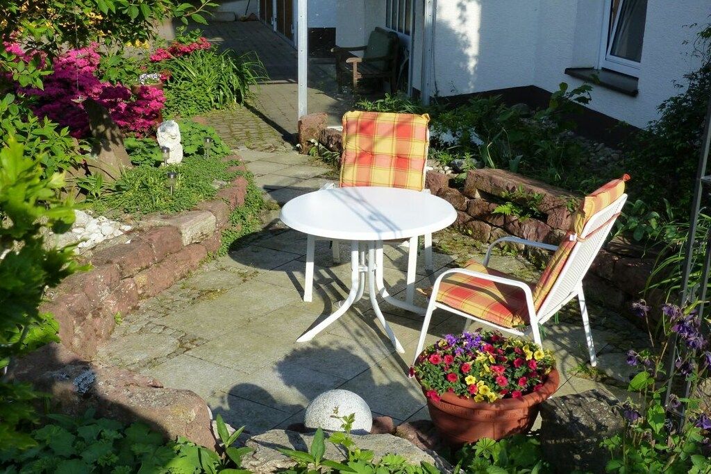Um pátio externo com mesa branca e duas cadeiras, cercado por um jardim com diversas plantas e flores.