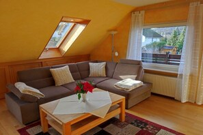 Ein Wohnzimmer mit einem braunen Sofa, einem Holztisch, einer Zimmerpflanze und einem Fenster mit Vorhängen.
