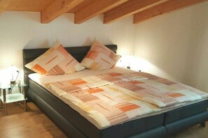 Uma cama com colcha estampada, duas almofadas, mesa de cabeceira com abajur e estrutura de teto em madeira.