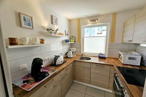 Une cuisine compacte avec des meubles en bois, un four à micro-ondes, une bouilloire et un grille-pain. On y trouve des étagères avec des tasses et un tableau encadré au mur.