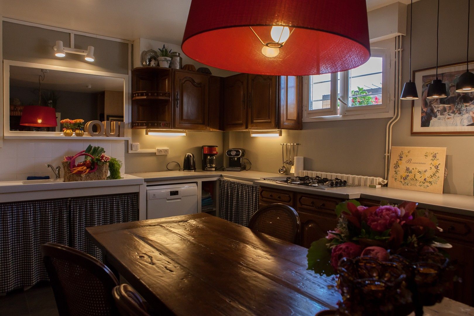 A kitchen with wooden cabinets, a red hanging light, a coffee machine, and a dining table with chairs.
