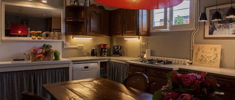 A kitchen with wooden cabinets, a red hanging light, a coffee machine, and a dining table with chairs.