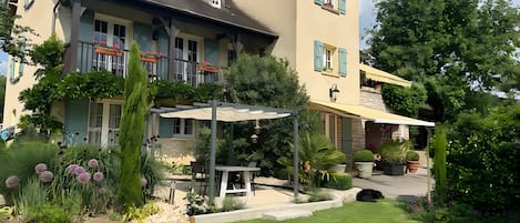 Ein zweistöckiges Gebäude mit überdachter Terrasse, einem kleinen Garten mit Kübelpflanzen und einer Sitzecke.