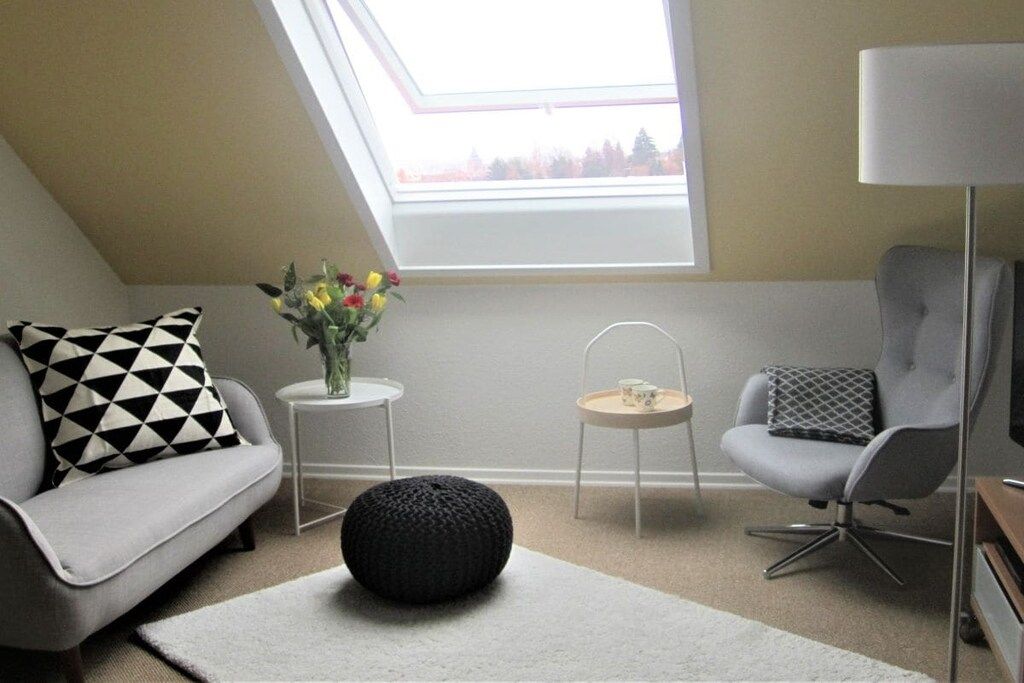 A modern living room with a sloped ceiling, a sofa, a chair, a small table with a vase of flowers, and a lamp.