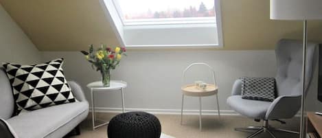 Una sala de estar moderna con techo inclinado, un sofá, una silla, una mesita con un florero y una lámpara.