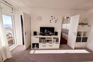 A room with a television on a white entertainment unit, a clock on the wall, and a view through an open door. - Holiday apartment with 1 bedroom (Sierksdorf)