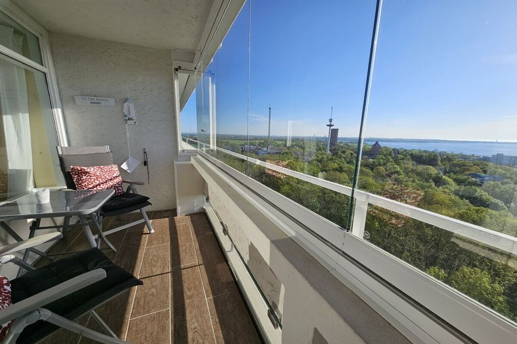 Ein Balkon mit Glasgeländer, der auf eine Stadtlandschaft und Grünflächen blickt.