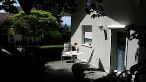 Um pátio externo com mesa e cadeiras, um vaso de planta e vista para um edifício com janela e porta.