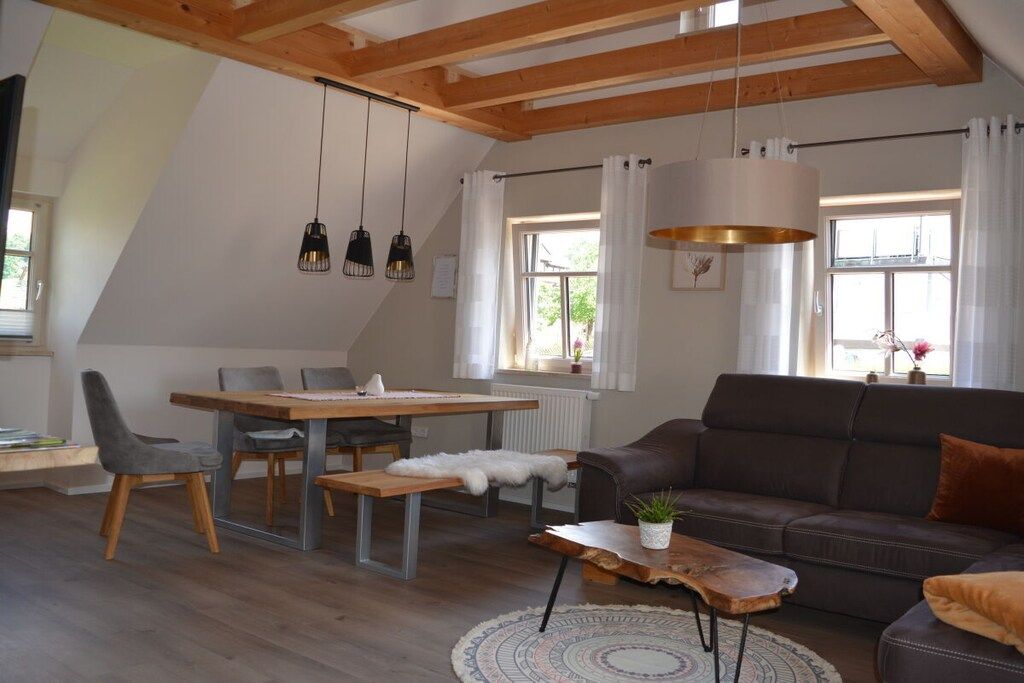 A modern living room with a dining area, wooden ceiling beams, pendant lights, and a sofa with a throw pillow.