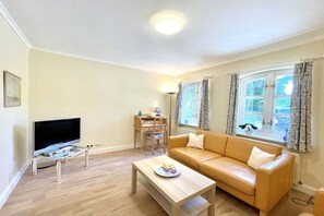 Een woonkamer met een bank, salontafel, televisie op een dressoir, bureau met stoel en een raam met gordijnen.