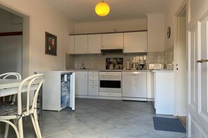 A compact kitchen with white cabinets, a built-in oven, and a small dining area with a white table and chairs. - Holiday apartment with 1 bedroom (Sierksdorf)