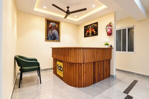 Zona de recepció d'hotel amb taulell de fusta, una cadira verda, ventiladors al sostre i obra d'art penjada a la paret.