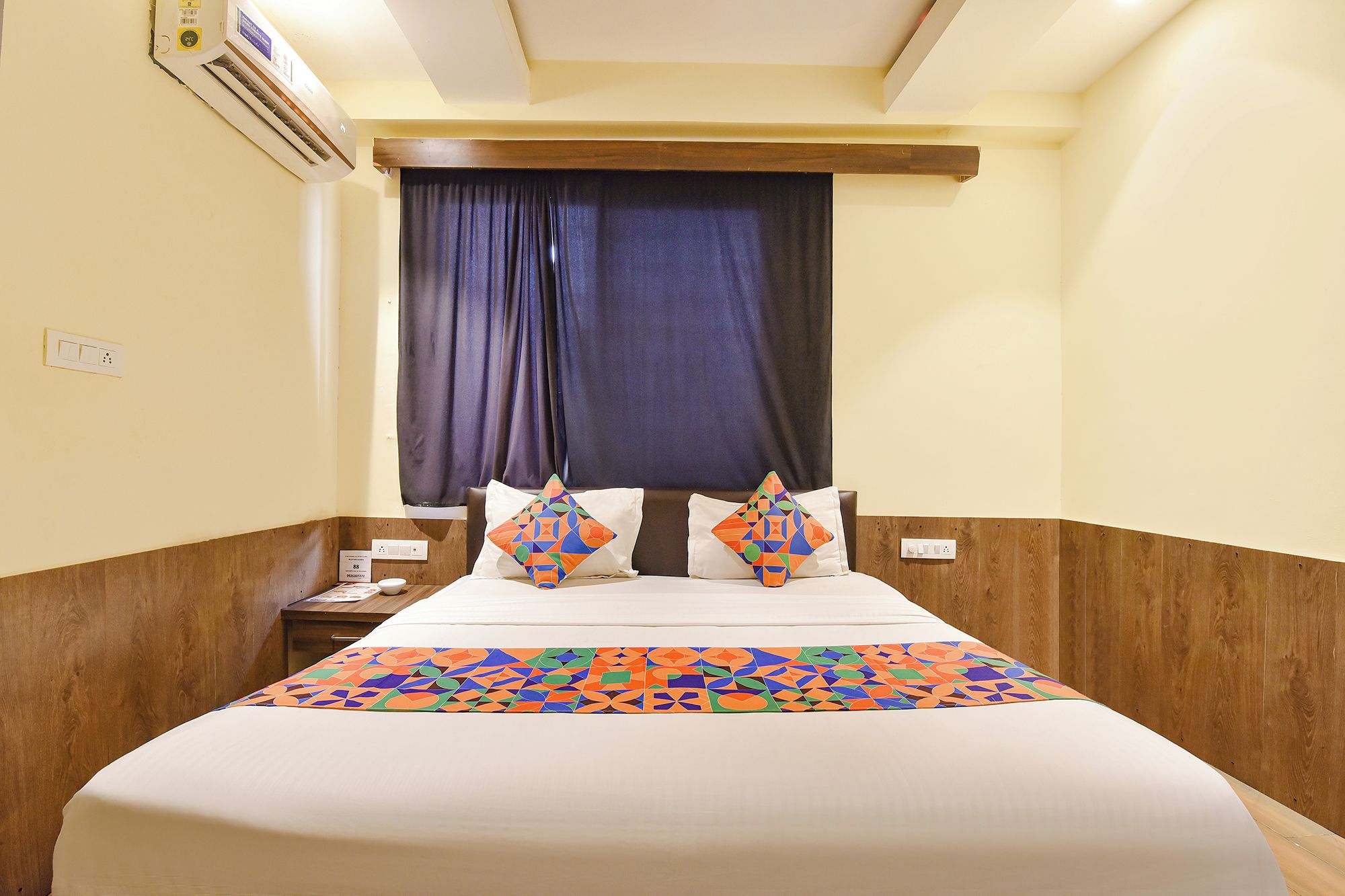 Quarto de hotel com cama, mesa de cabeceira, tomada e janela com cortinas.